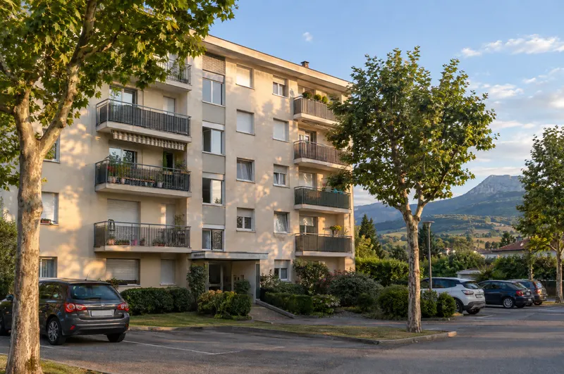 Façade d'un immeuble en copropriété avec balcons et parking arboré, en fin de journée dans le Nord-Isère